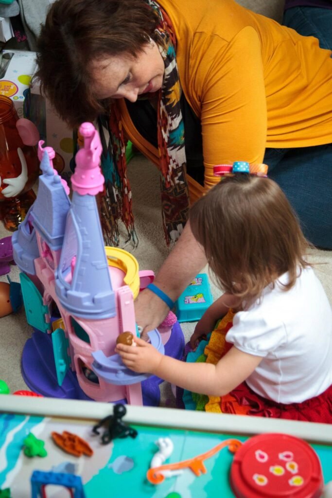 An adult and child play together with a colorful toy castle. A play table with various small items is visible in the foreground.