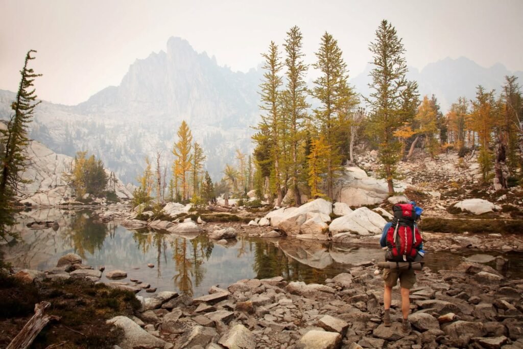 A hiker with a backpack stands on a rocky riverbank gazing at a calm waterway and tall evergreen trees.