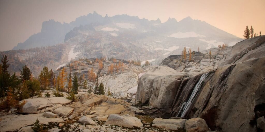 A wide landscape view of a small waterfall cascading over snow-dusted rocks, with evergreen trees and hazy mountains in the background.