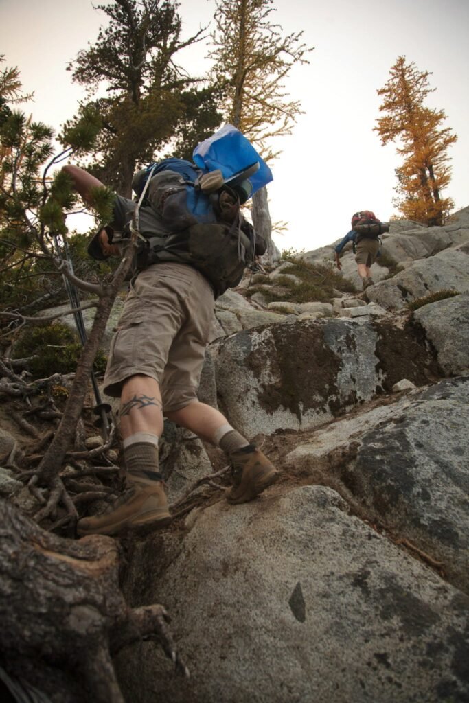 A hiker climbs a steep, rocky slope while another follows above against a backdrop of tall conifer trees.