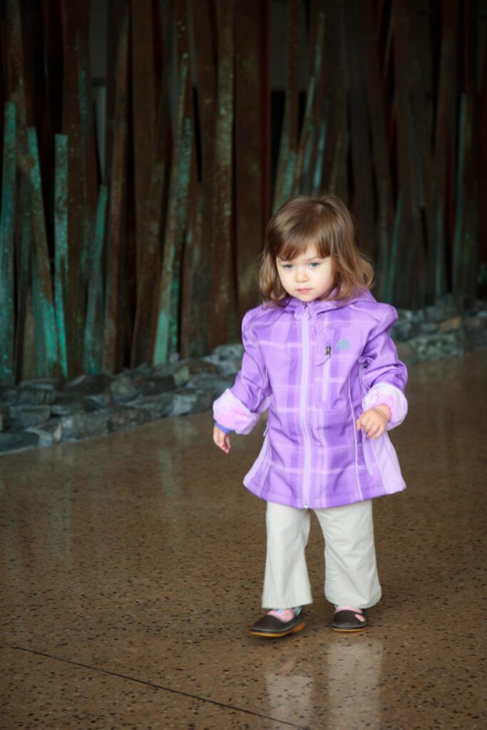 A child in a purple jacket and white pants standing in a public indoor space. The background features vertical architectural elements.