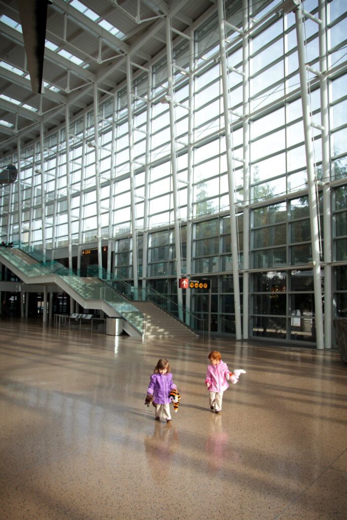 Two children walk through a massive, sunlit modern atrium with glass walls and long escalators.