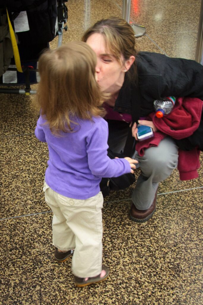 A person crouches down to kiss a young child on the forehead in a public indoor space. The child is wearing a purple top and white pants.
