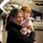 A smiling adult holds a small child with a stuffed bunny in a busy airport terminal. Background features modern architecture and other travelers.