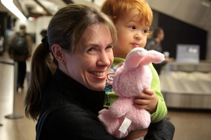 A smiling adult holds a small child with a stuffed bunny in a busy airport terminal. Background features modern architecture and other travelers.
