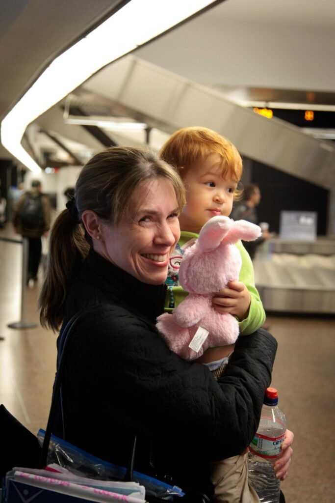 A smiling adult holds a small child with a stuffed bunny in a busy airport terminal. Background features modern architecture and other travelers.