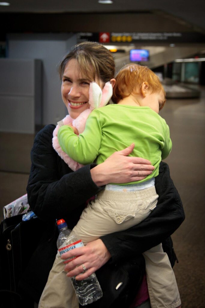 A person smiles while holding a child in a green outfit at an indoor public space, likely an airport, holding a water bottle.