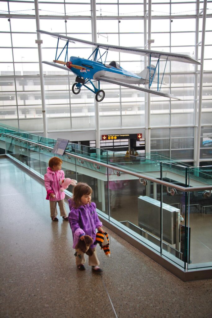 Children walk beneath a biplane model in a modern airport terminal.