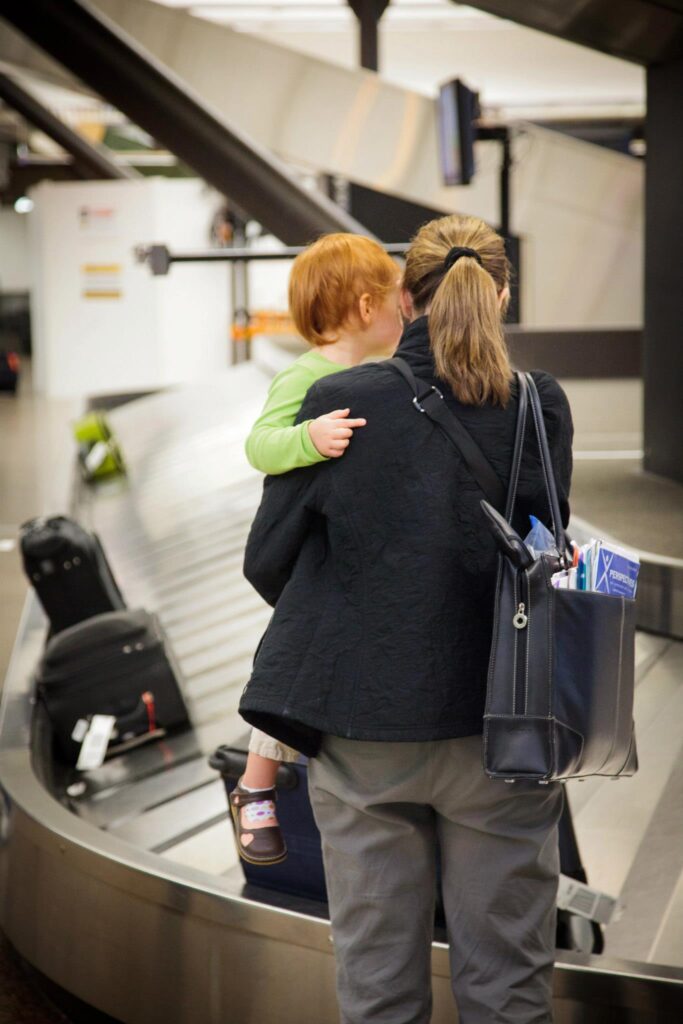 A person holds a child at an airport luggage carousel. Suitcases are visible on the moving belt in a busy terminal.
