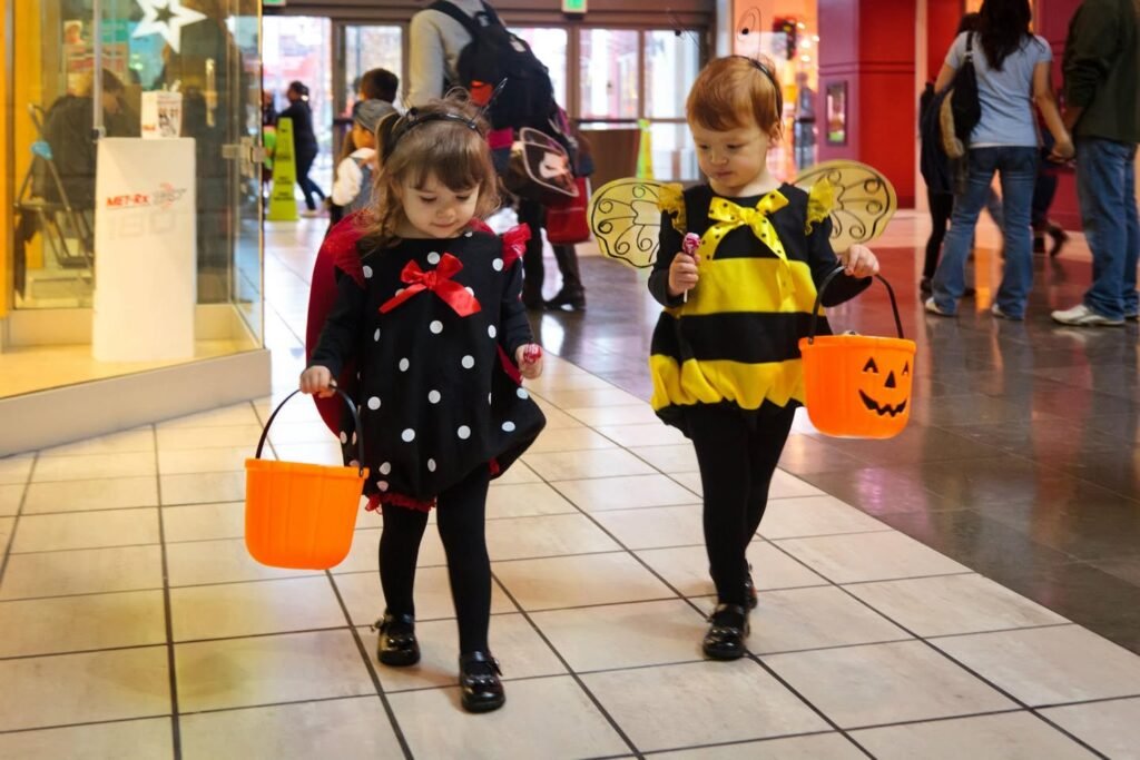 Two children in Halloween costumes walk through a shopping mall. One wears a polka-dot dress, the other a bee outfit, each carrying a jack-o'-lantern bucket.