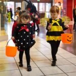 Thanksgiving at Cannon Beach Two children in Halloween costumes walk through a shopping mall. One wears a polka-dot dress, the other a bee outfit, each carrying a jack-o'-lantern bucket.