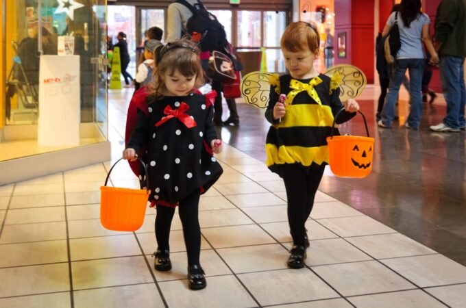 Two children in Halloween costumes walk through a shopping mall. One wears a polka-dot dress, the other a bee outfit, each carrying a jack-o'-lantern bucket.