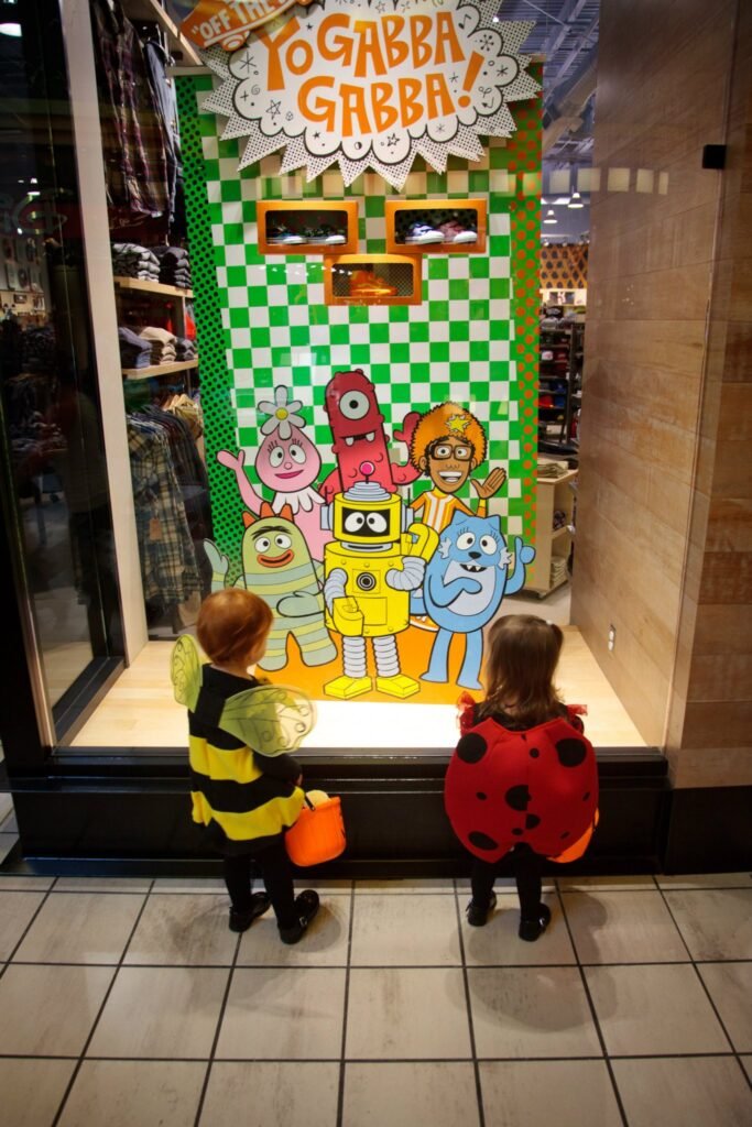 Two children in insect costumes stand before a colorful Yo Gabba Gabba themed store window display featuring cartoon characters.