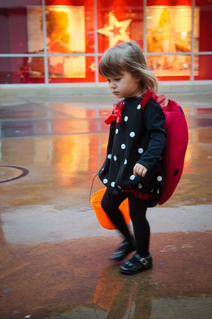 A child in a polka dot coat and red backpack walks across a wet plaza in front of a festive red display.