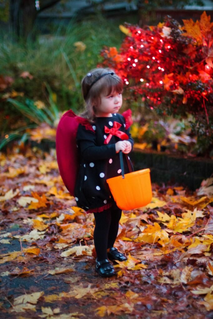 A young child in a witch costume holding an orange bucket amidst autumn foliage and red decorative bush.
