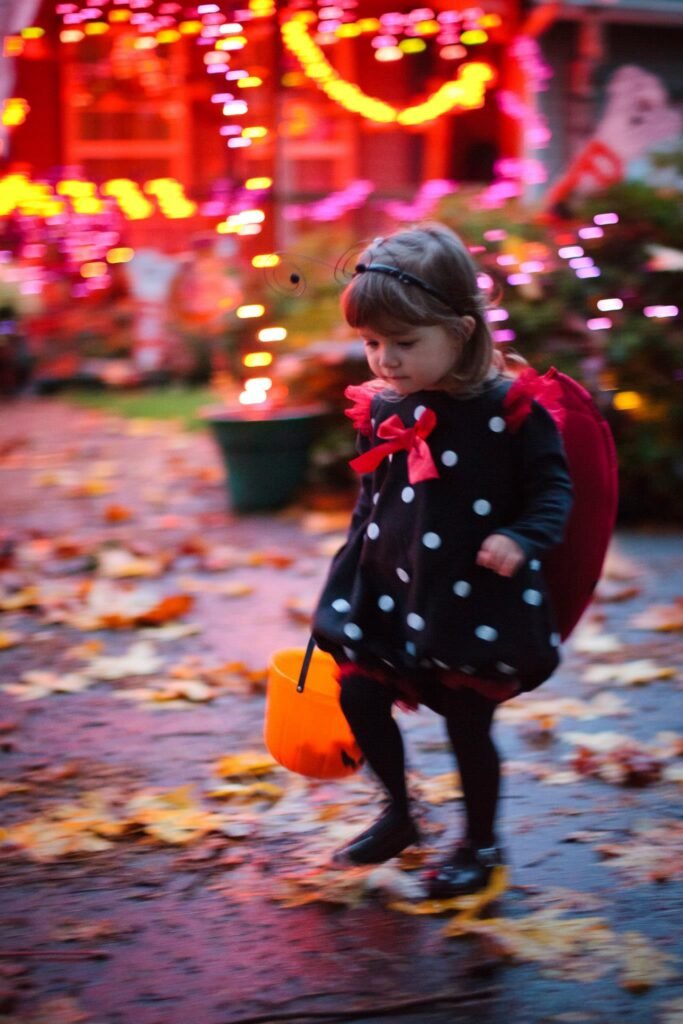 A child in a black polka dot dress and red cape walks a leaf-covered path at night. The background is bright with festive orange and pink holiday lights.