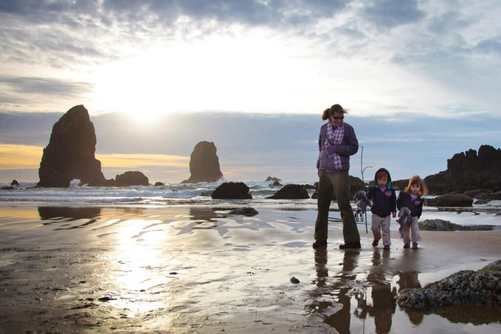 A family strolls across a reflective beach at sunset near rock sea stacks while the sun sets over the ocean.