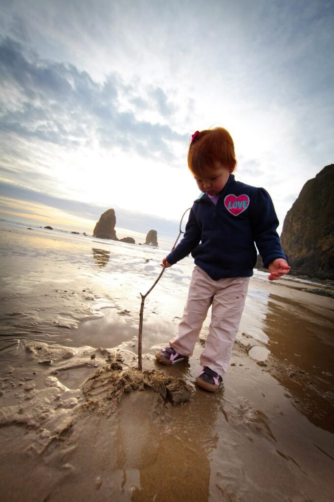 A child digs in the sand of a wet Pacific Northwest beach with rock formations in the background under a cloudy sky.