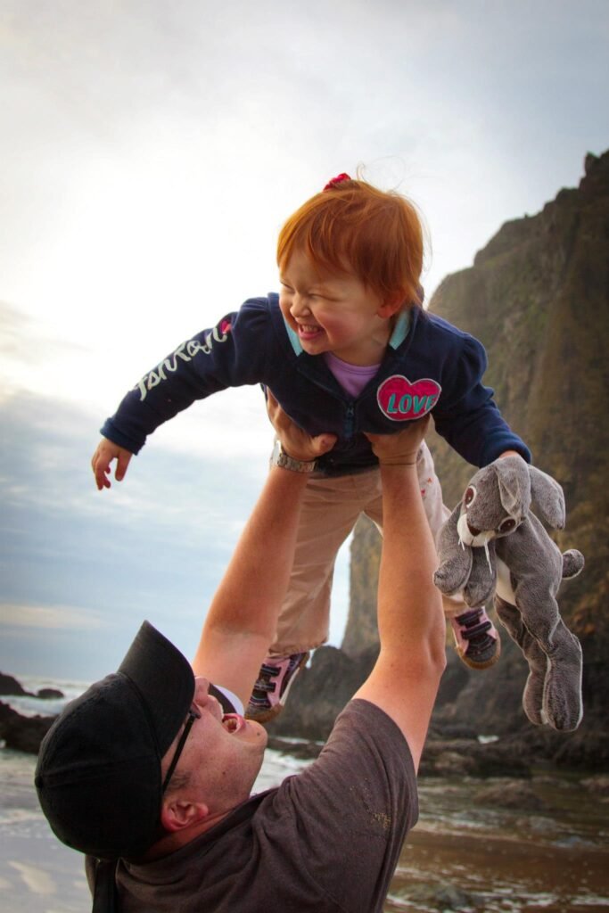 A happy child is lifted into the air while holding a stuffed bunny. The family enjoys a day together at the rocky Pacific Northwest shoreline.