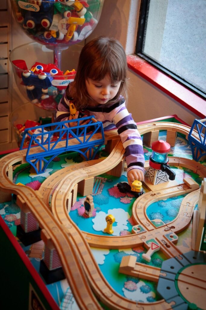 A child in a striped sweater playing with a wooden train set near a colorful toy gumball machine.