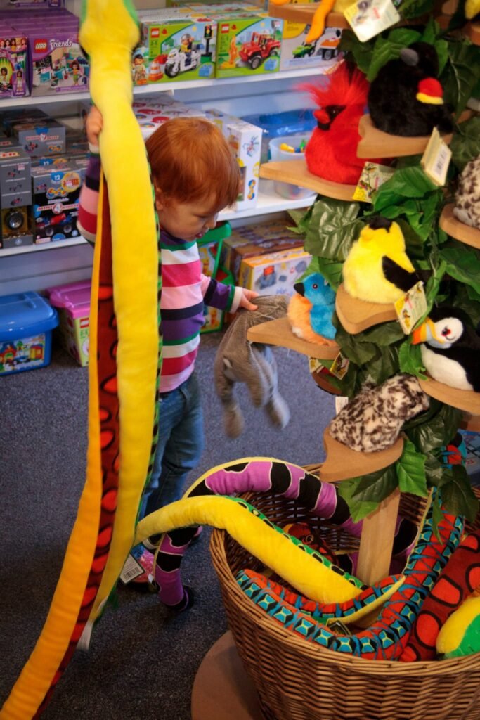 A child holds a long plush snake toy near a display of stuffed animals in a store. A basket filled with colorful toys sits in the foreground.