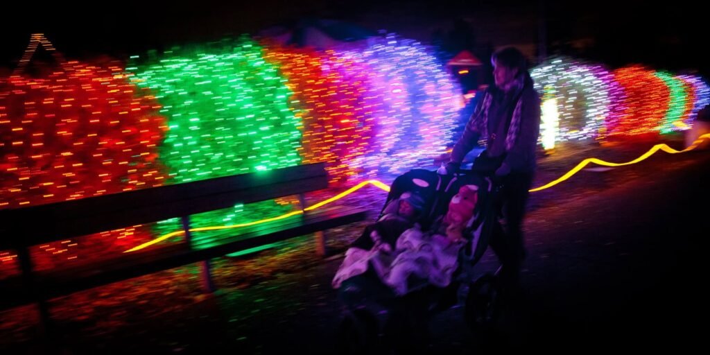 A family with a stroller stands in front of colorful holiday lights at night. The vibrant display creates a festive atmosphere.