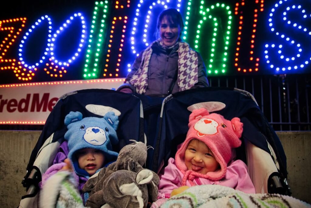 Children in colorful coats sitting in a stroller with a zoo lights sign behind them.