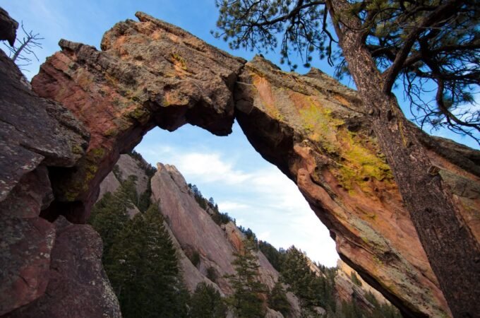 A natural stone arch frames a mountain landscape with evergreen trees below, set against a blue sky.
