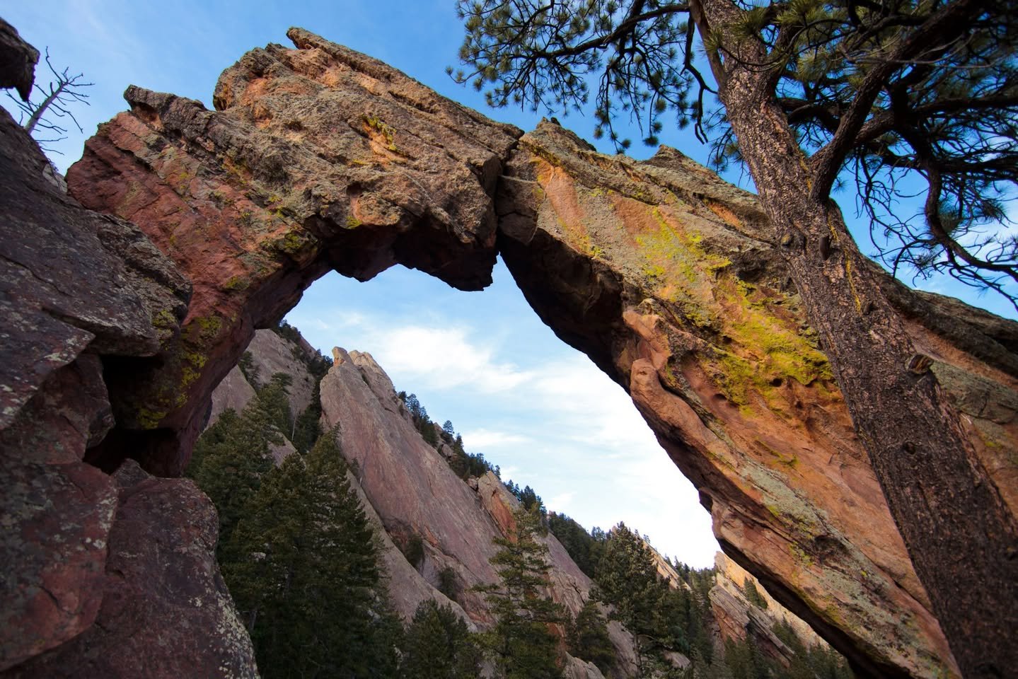 Royal Arch A natural stone arch frames a mountain landscape with evergreen trees below, set against a blue sky.