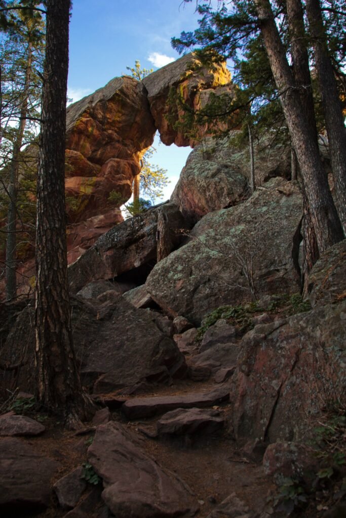 A large natural rock arch stands among tall pine trees with a clear blue sky in the background. The rugged terrain features rough stone surfaces and scattered greenery.