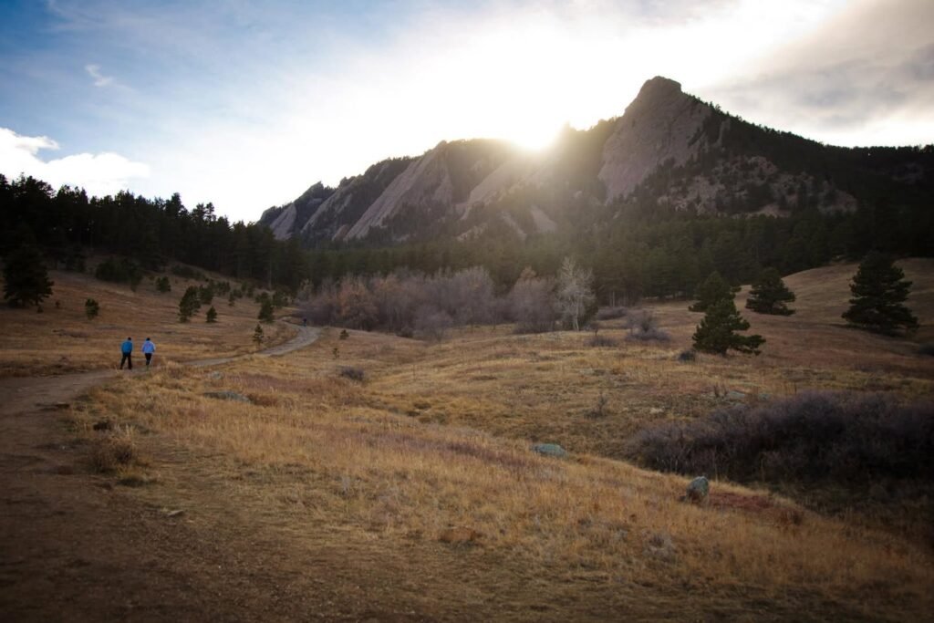A panoramic view of golden grasslands and a rocky mountain peak at sunrise. The sun shines brightly from behind the ridge, casting a warm glow over the landscape.