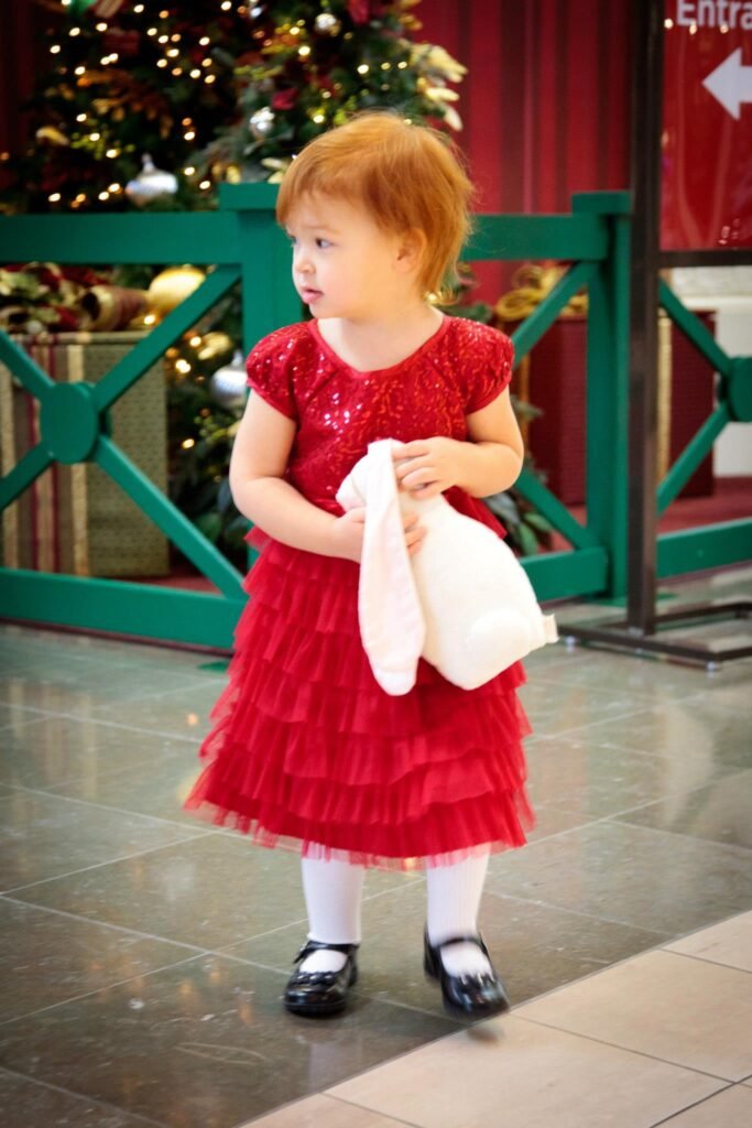 A toddler in a red dress stands near a green railing in front of a Christmas tree.