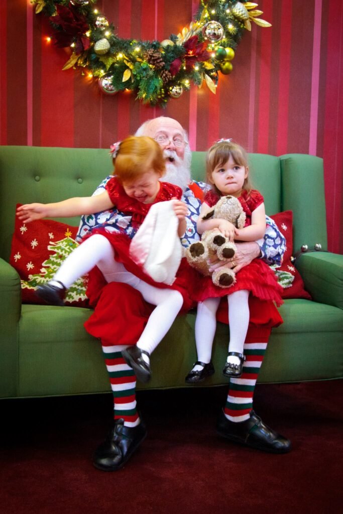 A group of children in festive red dresses sit on a green couch with a Christmas wreath in the background.