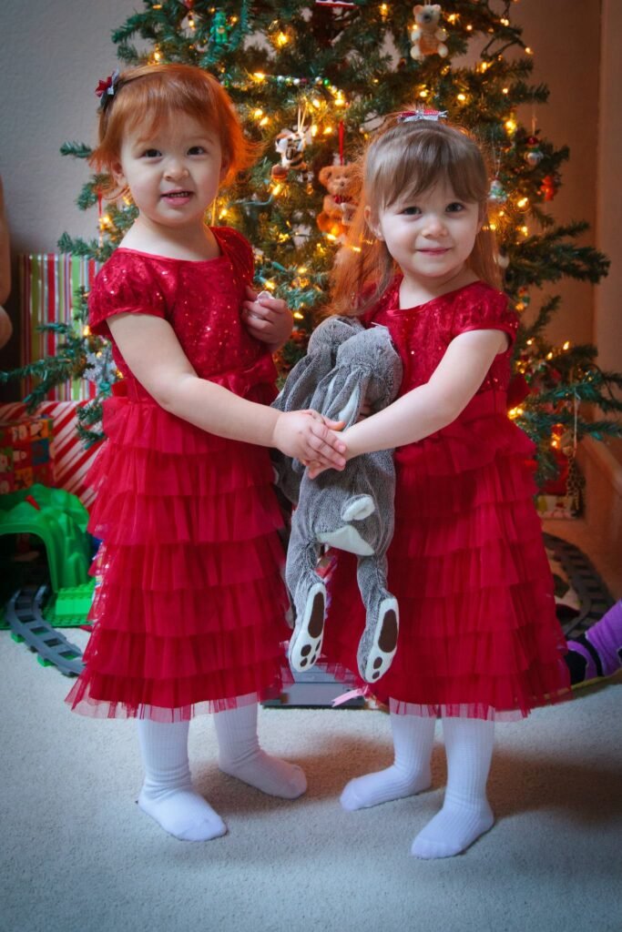 Two children in red dresses standing under a lit Christmas tree. They share a grey stuffed animal and white tights.