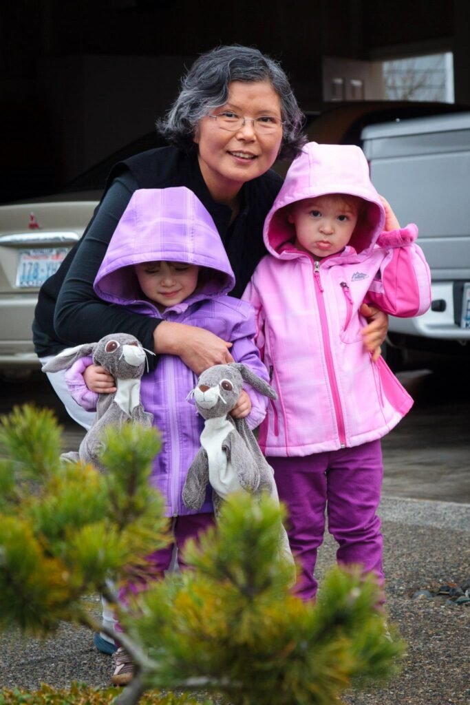 A joyful family moment in the rain, featuring a woman and two children in colorful raincoats. The kids are holding stuffed bunnies, surrounded by a cozy, rainy suburban setting.