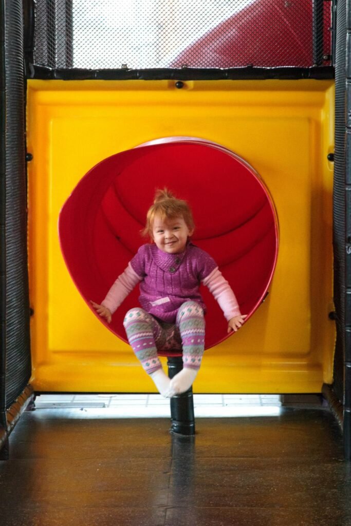 A smiling toddler sits inside a brightly colored red circular structure against a yellow wall. This indoor play area features a polished wooden floor and a concrete exterior.