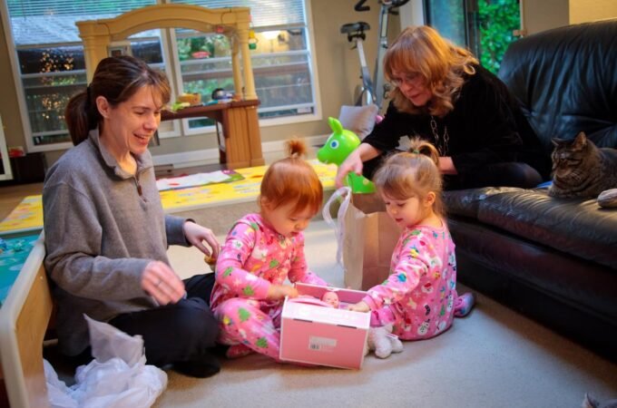 A family is gathered in their home while two children open a pink gift box on the floor.