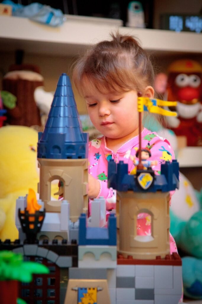 A child wearing pink pajamas plays with a disney-style princess castle toy set, featuring blue roofs and a drawbridge, surrounded by stuffed animals.