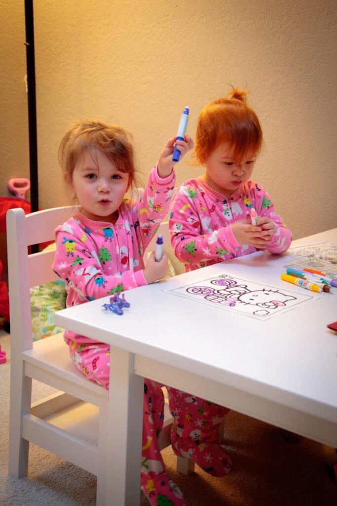 Children sitting at a white table coloring a piece of paper with markers at home.