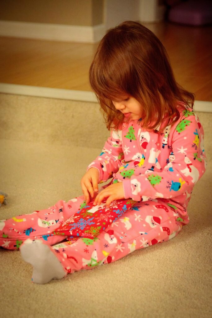 A young child in pink Christmas pajamas sits on a carpeted floor opening a gift. Wearing patterned flannel pajamas with festive designs and white socks, looking down with anticipation.