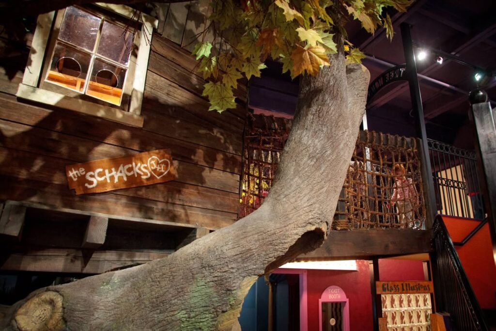 A dinosaur exhibit features a standing skeleton next to a wooden facade with a window. Yellow artificial leaves hang from a large branch in a dimly lit room.