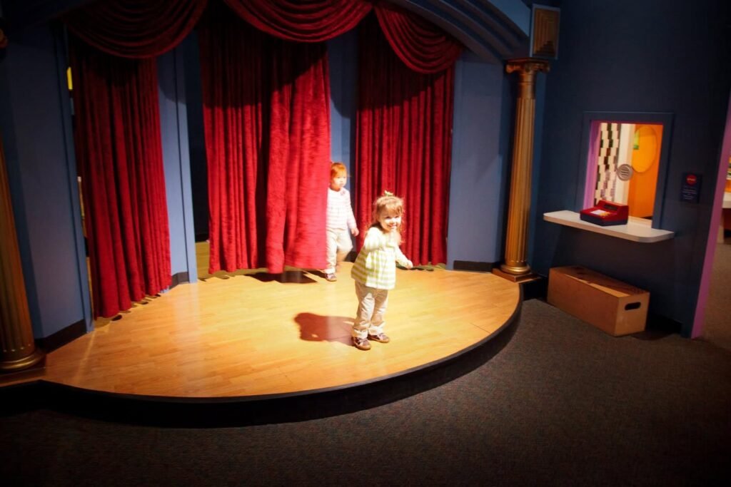 Two small children stand on a wooden stage in front of red curtains with a framed artwork on the side.