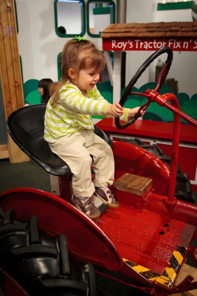 A young child sitting in a red toy tractor in a play area, holding the steering wheel.