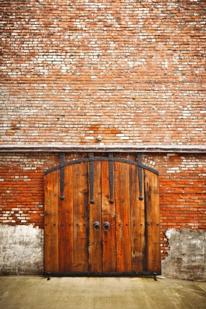 A vibrant orange-red brick wall with weathered patches and a heavy wooden door. Close-up details highlight the rustic texture and architectural durability of the structure.