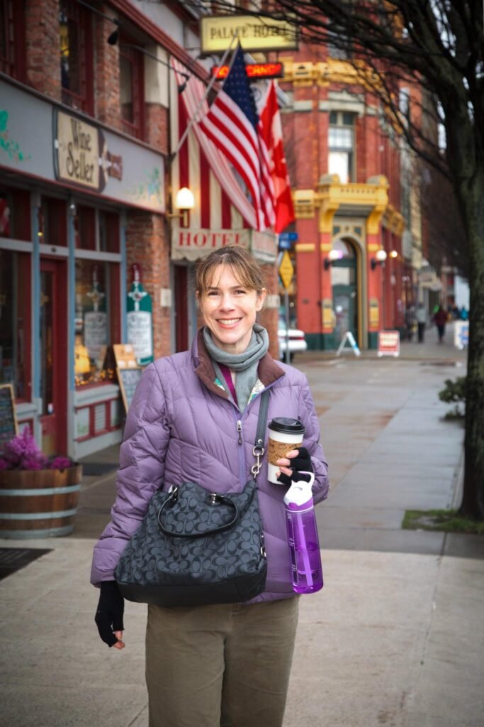 A person walks along a historic street in Vancouver, British Columbia, holding a coffee cup and a purple bottle. Red brick buildings and an American flag are visible in the background.