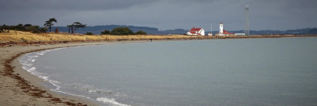 A scenic view of a lighthouse on a distant shore with a sandy beach and calm waters in the foreground.