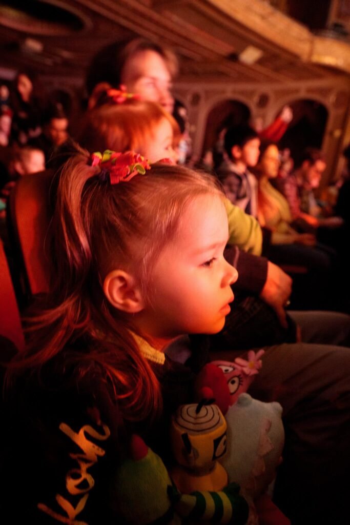 A child watches a performance in a dimly lit theater. The audience is visible in the background.