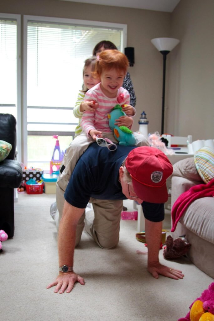 A playful living room scene in a cozy home. A child sits on a person's back while another child stands nearby.