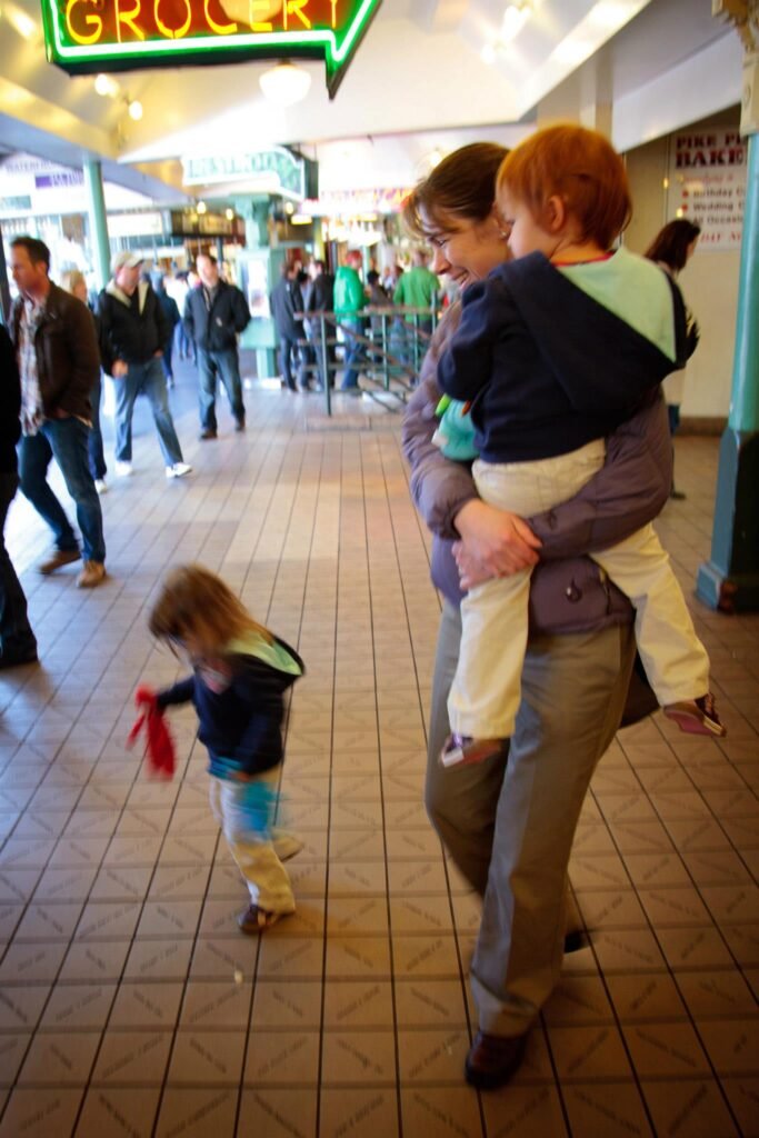A parent carrying a child and a toddler walking in a busy airport terminal under a neon sign.