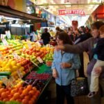 A group of people browsing a colorful fruit stand in a bustling indoor market under a 'Lower Floor' sign.